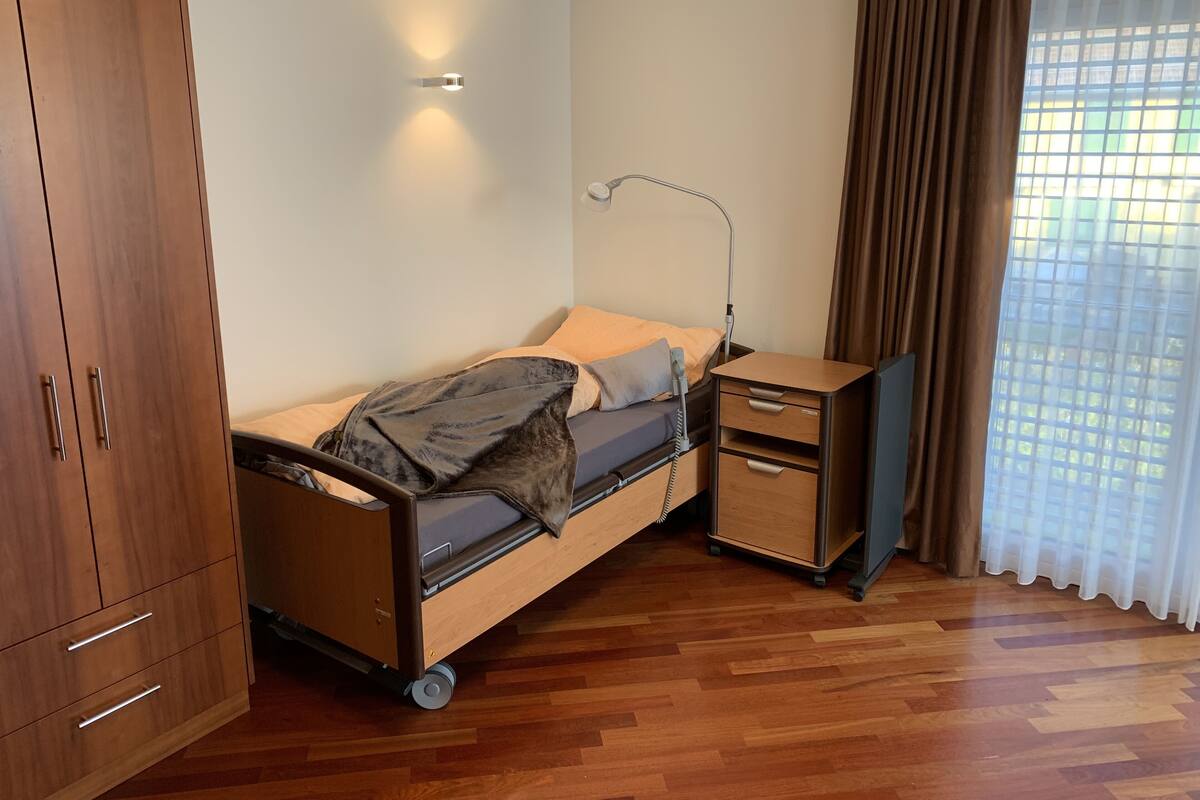 A single-bed room with a wooden floor, a bedside table with drawers, a lamp, and a window with curtains.