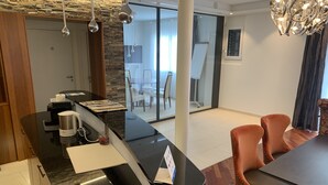 Une cuisine moderne avec un plan de travail noir, une machine à café et une bouilloire. On y trouve également un coin repas avec une table et des chaises.