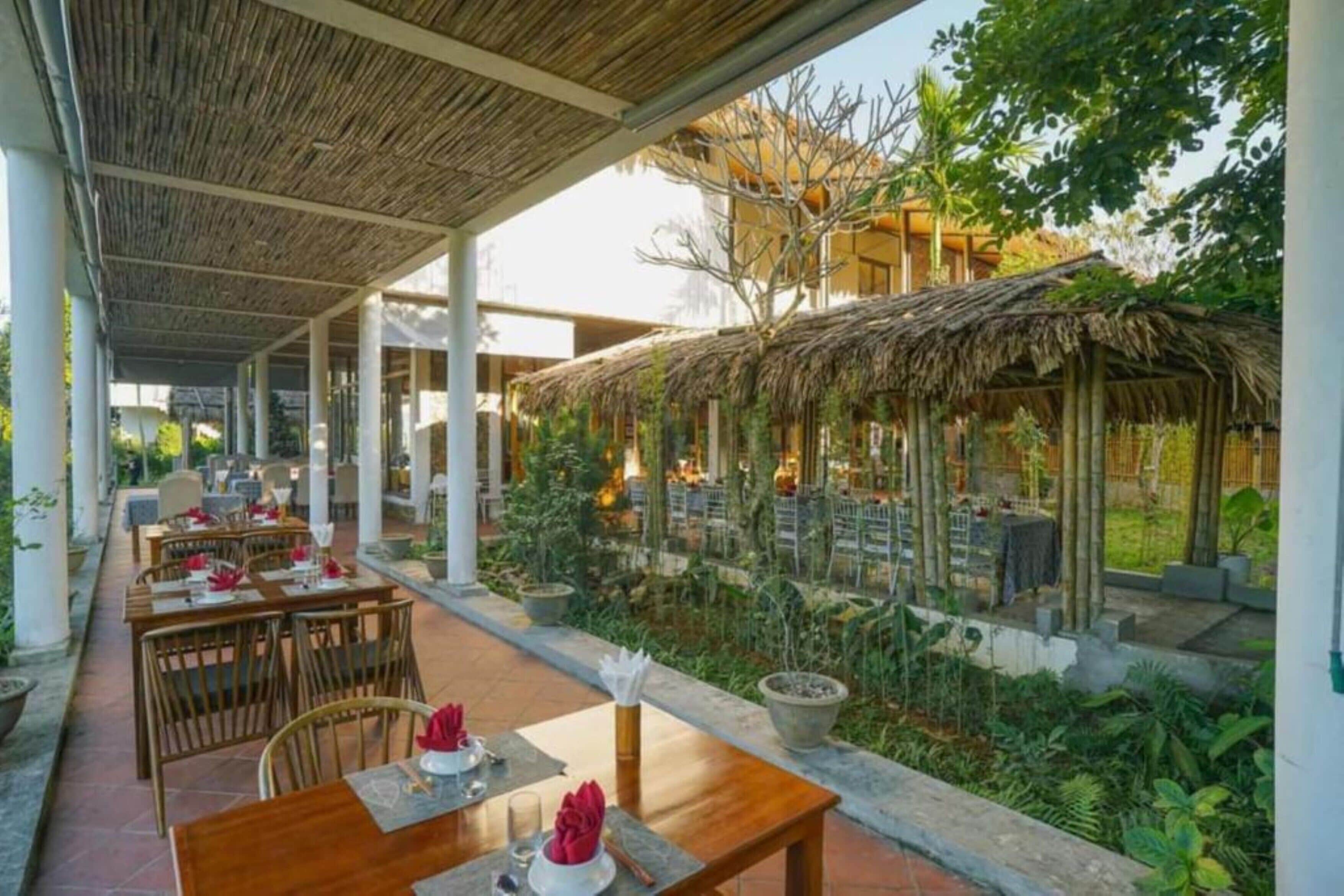 Un espace de restauration en plein air, avec des tables et des chaises, entouré de verdure et abrité par une structure à toit de chaume.