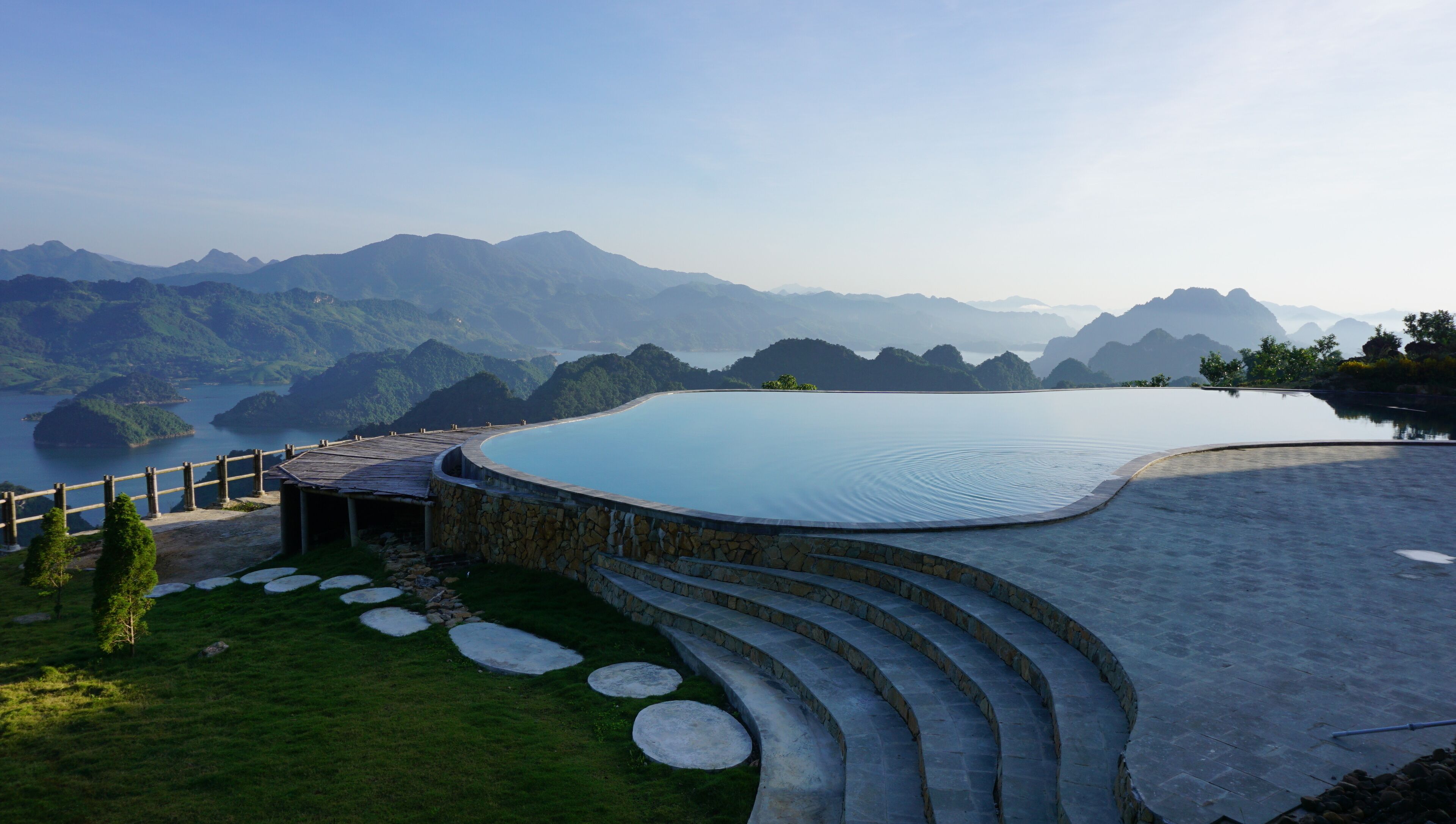 A large, curved infinity pool with a stone deck and steps leading into the water, surrounded by lush greenery and mountains in the background.