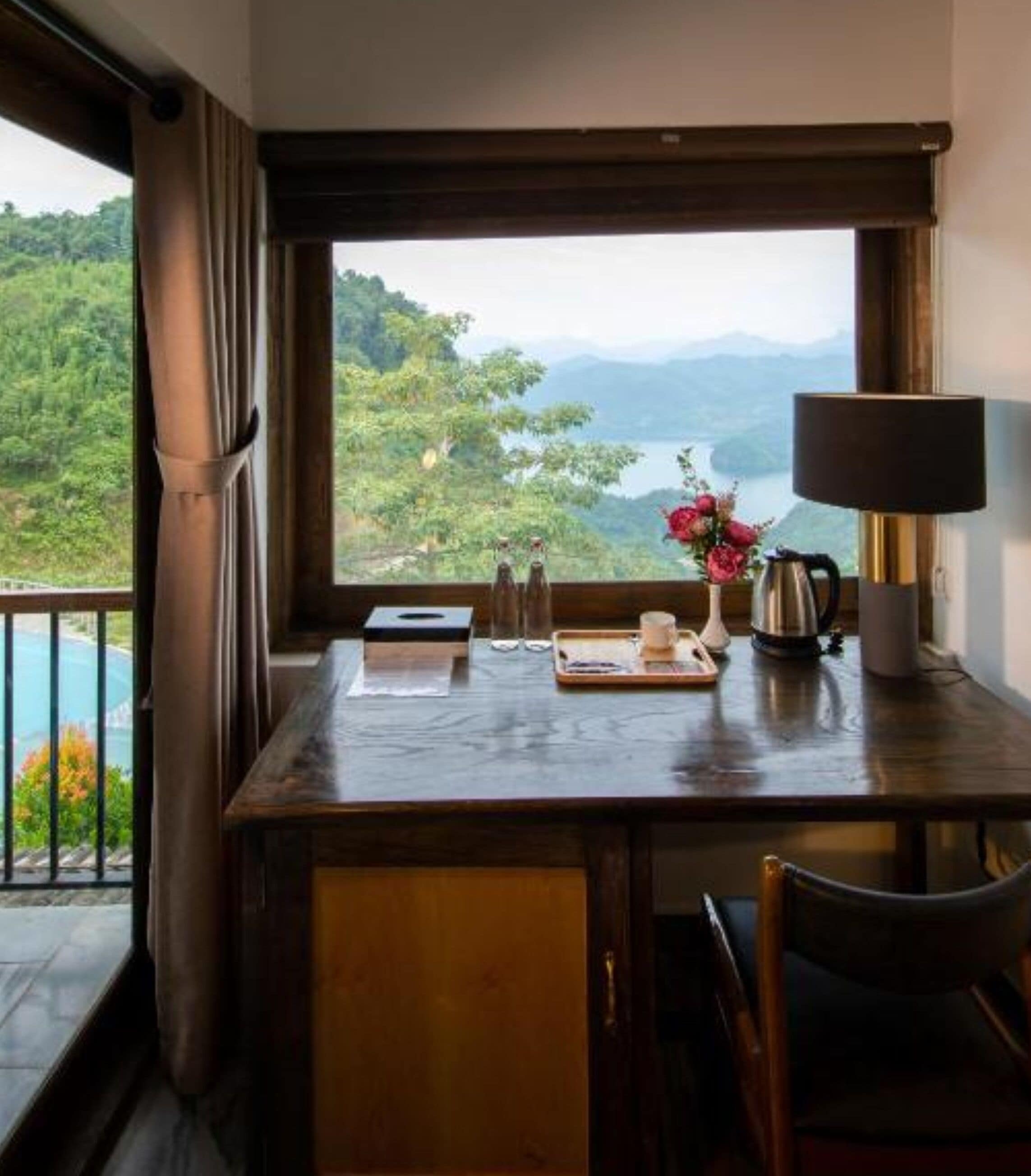 A wooden table with a view of a mountainous landscape through a window, a lamp, and a tray with a cup and a teapot.