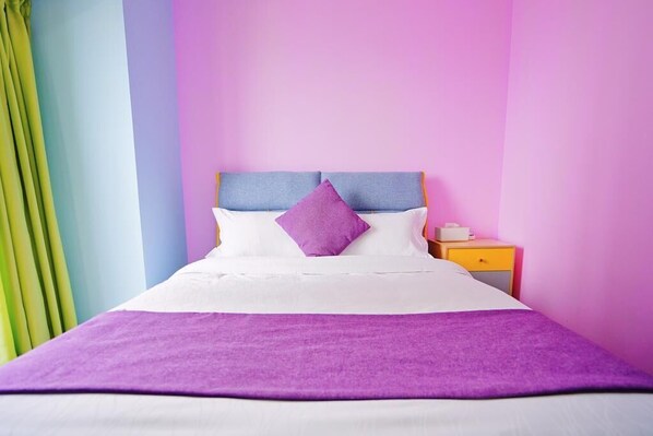 Un lit recouvert d’une couvre-lit blanche et violette, avec une tête de lit bleue et un oreiller violet. On y trouve une table de chevet avec une lampe blanche et un tiroir jaune.