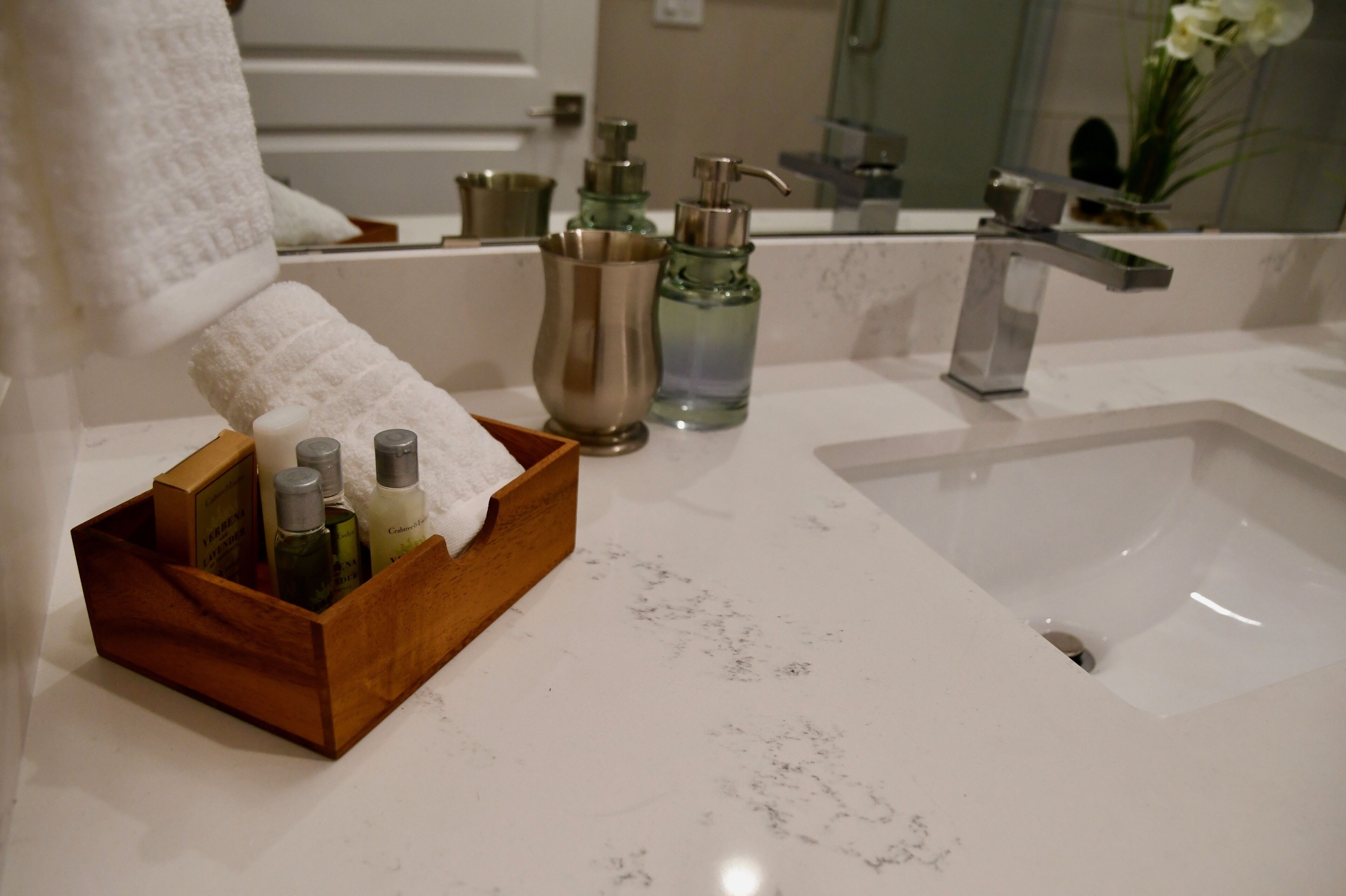 A bathroom with a marble sink, a wooden tray holding toiletries, and a mirror reflecting a vase with flowers.