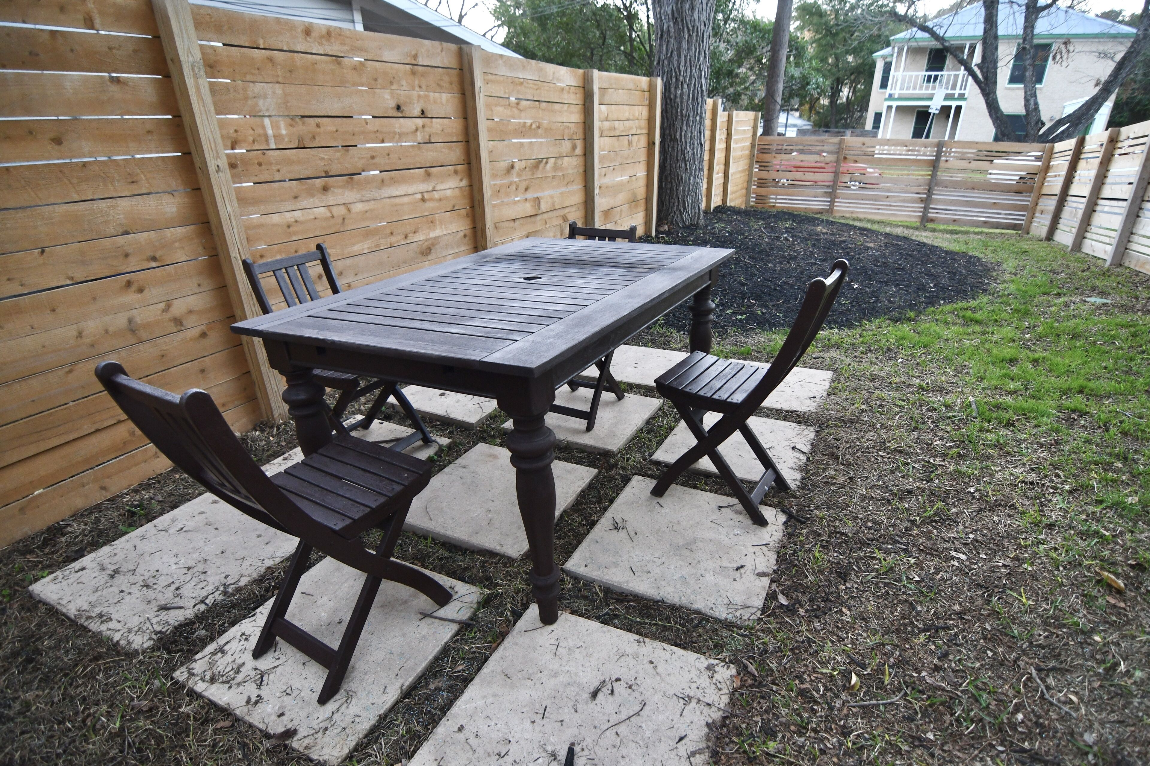 A wooden table and chairs set on a stone patio with a wooden fence in the background.