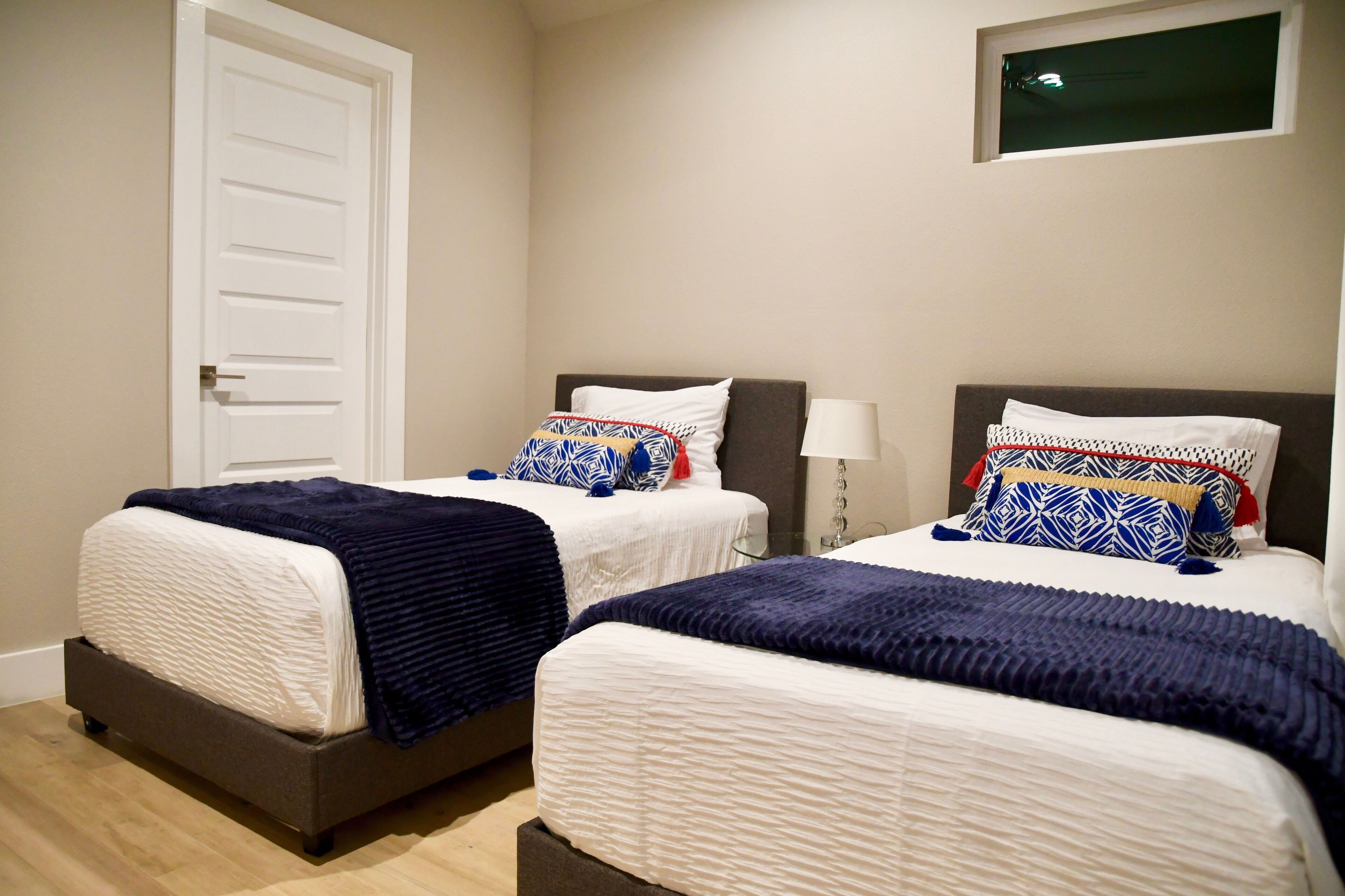 A hotel room with two beds, each with a white mattress and dark blue bedding. There is a white door on the left and a window above the beds.