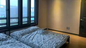 A bedroom with two beds, large windows, a door, and a view of buildings outside.