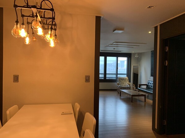 A modern dining area with a white table and chairs, a hanging light fixture, and a large window offering a view of the city.