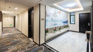 A hotel corridor with a patterned carpet, a mural with a boat and mountains, potted plants, and a modern ceiling design. - Yiwu Luck Bear Hotel (Jinhua)