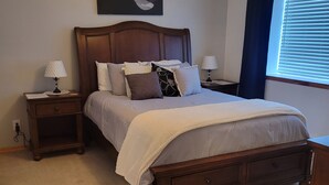 A bedroom with a wooden bed, two bedside tables with lamps, a ceiling fan, a window with blinds, and a wall-mounted artwork. - Shaggy Moos Apartment  (Bloomfield)