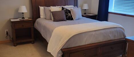 A bedroom with a wooden bed, two bedside tables with lamps, a ceiling fan, a window with blinds, and a wall-mounted artwork.
