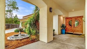 A house entrance with a stone fountain, a wooden double door, and a landscaped garden. - Central Coast Casa II (Atascadero)