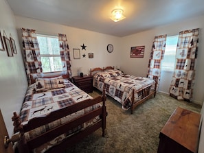 Quarto com duas camas de madeira, cortinas xadrez, um relógio de parede e uma bandeira americana emoldurada.