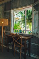 Una mesa de madera con sillas, una lámpara de pie de mimbre y una ventana con vistas a plantas verdes.