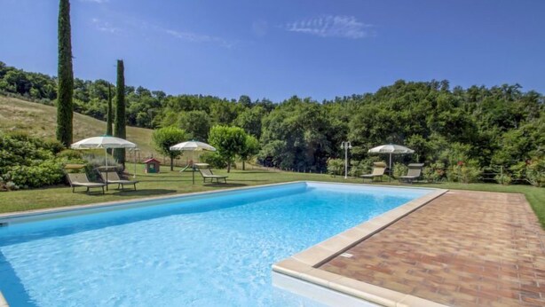 A rectangular swimming pool with a tiled edge, surrounded by a brick patio. There are lounge chairs and umbrellas around the pool area.