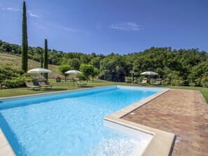 Uma piscina retangular com borda revestida, rodeada por um pátio de tijolos. Há espreguiçadeiras e guarda-sóis na área da piscina.