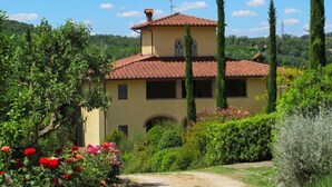 A two-story building with a tiled roof, surrounded by lush greenery and tall cypress trees.