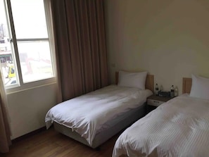 A hotel room with two beds, a window with curtains, a bedside table with a lamp, and a view of the street outside. - Old Harbour Hostel (Nangan)