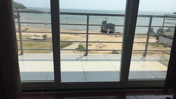Una habitación con un amplio ventanal con vista a una playa, suelo de hormigón y barandilla de metal.