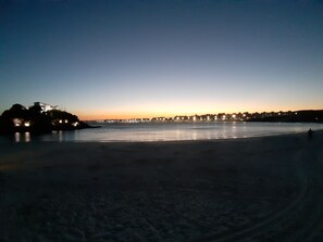 Uma praia ao entardecer com um horizonte urbano distante e uma ponte.