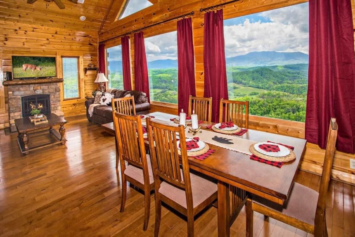 A wooden cabin interior with a dining table set for four, a fireplace, and a large window overlooking a mountainous landscape.