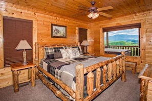 A wooden cabin bedroom with a bed, two bedside lamps, a ceiling fan, a painting, and a window with a view of green hills. - Stunning View | Hot Tub & Resort Pool Access (Sevierville)