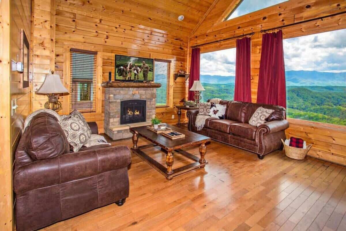 A log cabin interior with a fireplace, leather sofas, wooden flooring, and a large window with a mountain view.