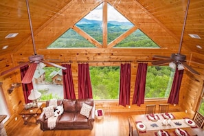 A wooden cabin interior with a large window, ceiling fans, a sofa, and a dining table. - Stunning View | Hot Tub & Resort Pool Access (Sevierville)
