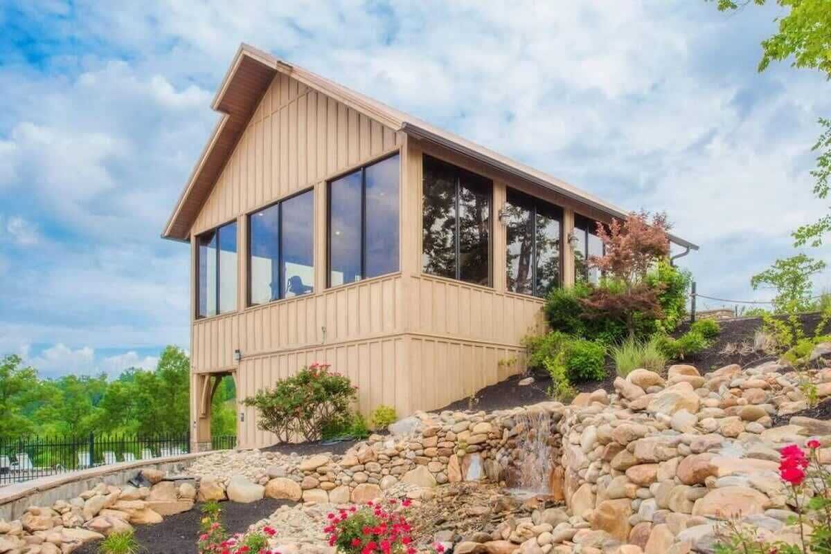 A modern wooden building with large windows, a stone waterfall feature, and a garden with rocks and flowers.