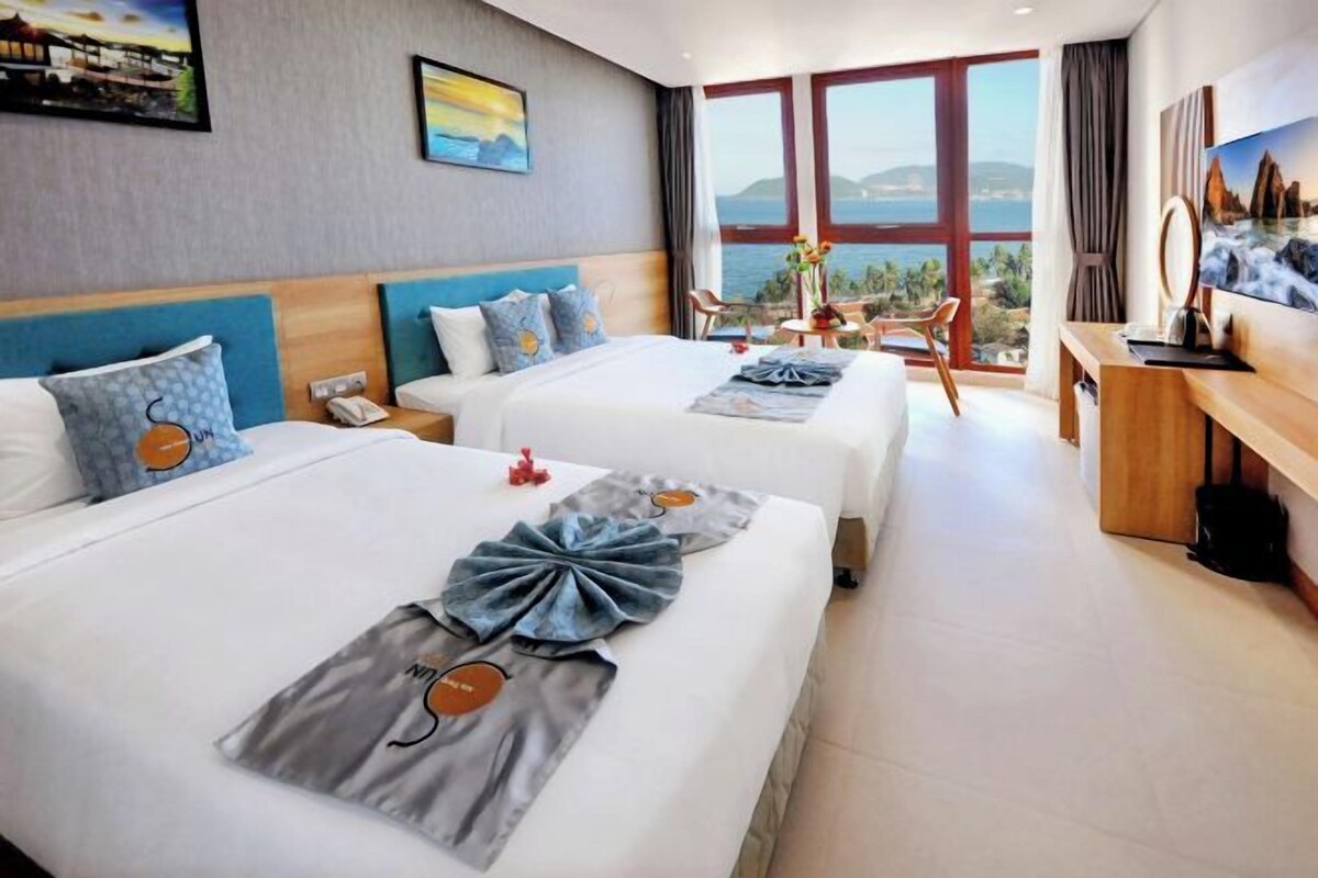 A hotel room with two beds, a desk, and a view of the sea.
