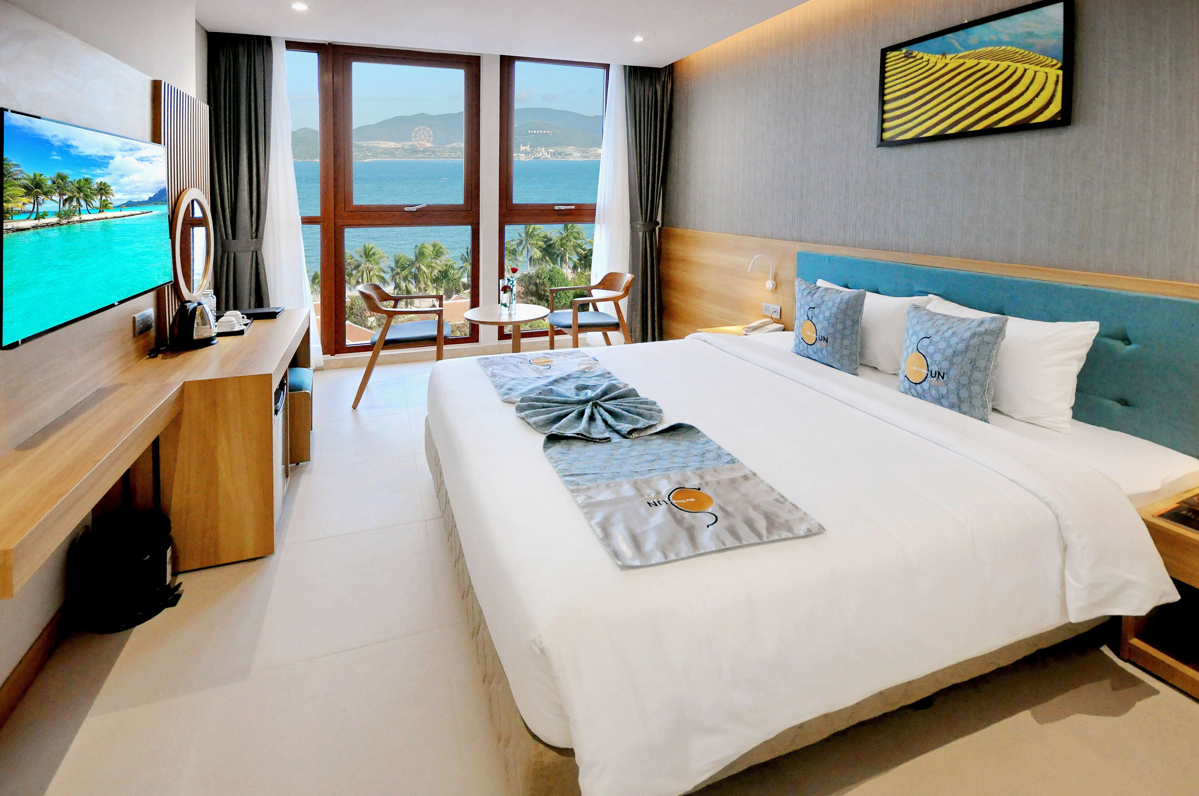 Ein Hotelzimmer mit einem großen Bett, einem Fernseher links, einem Schreibtisch mit Stuhl und Blick auf das Meer durch große Fenster.