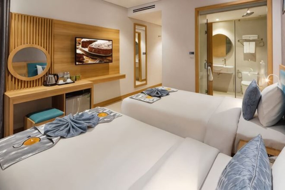 Habitación de hotel con dos camas, un escritorio, un espejo, un televisor y un baño visible a través de una puerta abierta.