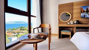 A hotel room with a large window offering a view of the sea and a distant mountain range. There is a wooden desk with a round mirror, a bed, and a small table with a magazine. - Sunkiss Hotel (Nha Trang)