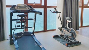 Una habitació àmplia amb grans finestres que donen a una massa d'aigua, equipada amb cinta de córrer i bicicleta d'exercici.