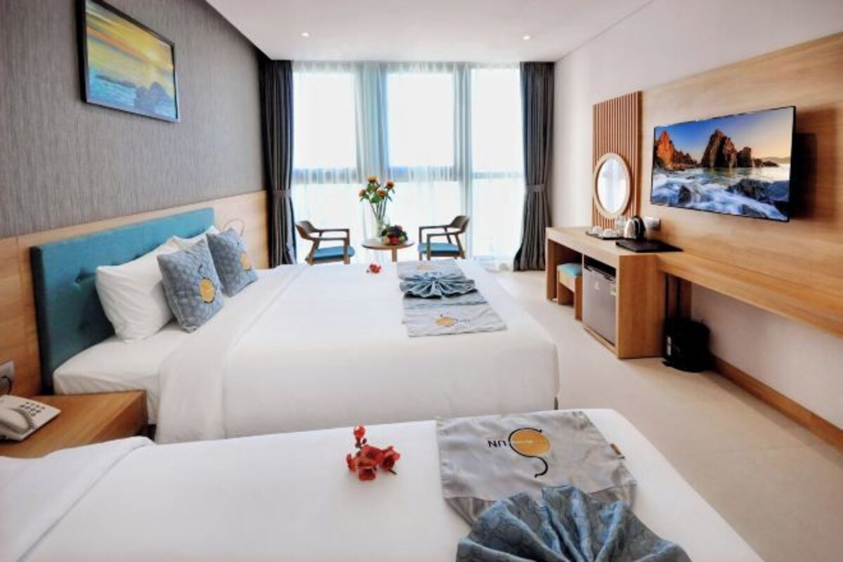Habitación de hotel con dos camas, un televisor, un escritorio con silla y vistas al mar a través de amplias ventanas.