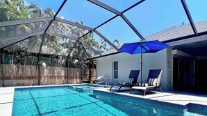 A swimming pool with clear water, surrounded by a covered area with lounge chairs and an umbrella. - 555 Royal Palm Villa | Luxury Heated Pool Home (Naples)