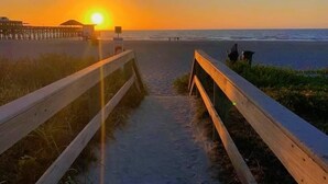 Een houten looppad dat naar een strand leidt, met een ondergaande zon op de achtergrond.