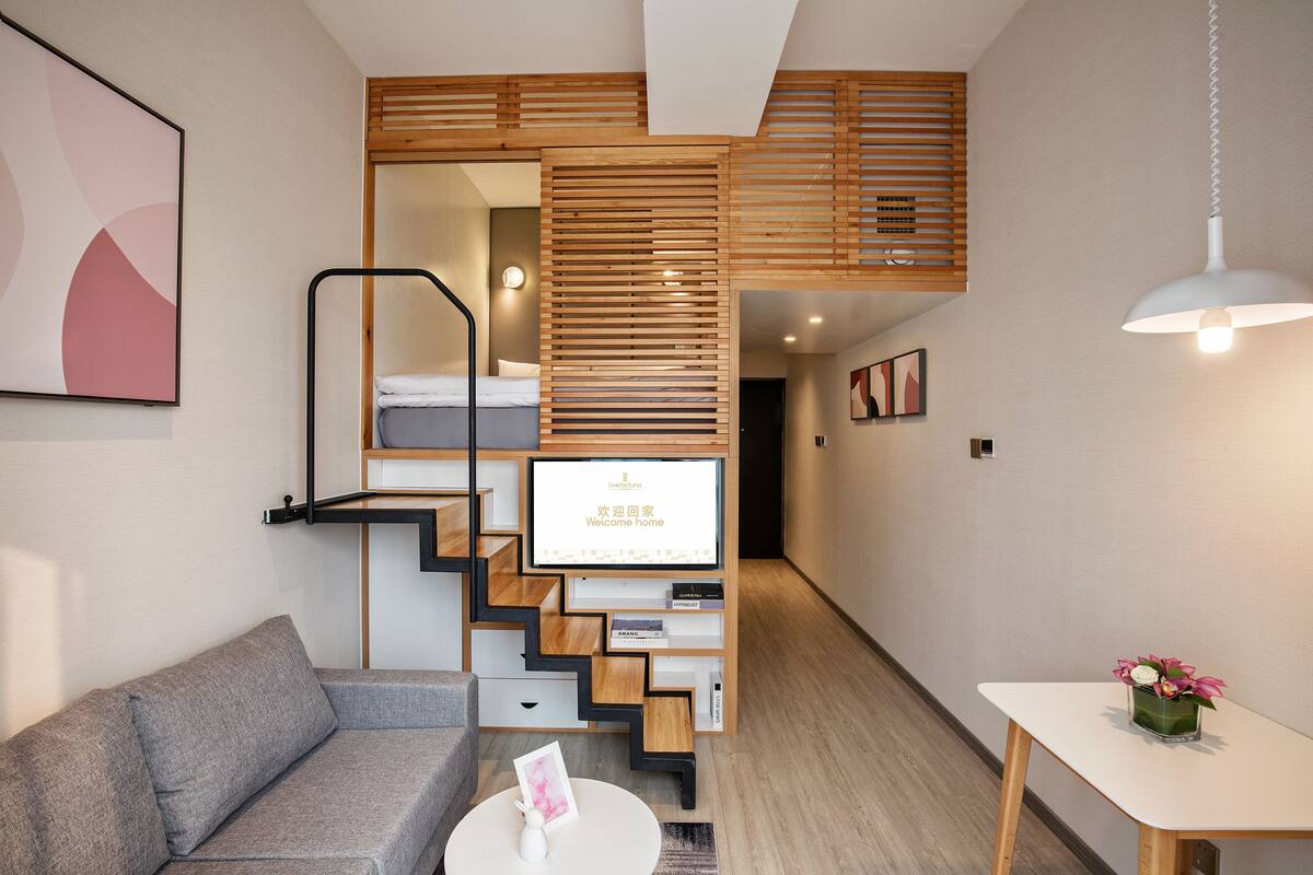 Ein modernes Hotelzimmer mit einer Couch, einem kleinen Tisch mit einer Vase, einem Fernseher, der 'Willkommen Zuhause' anzeigt, und einer Treppe, die zu einem Schlafzimmer führt.