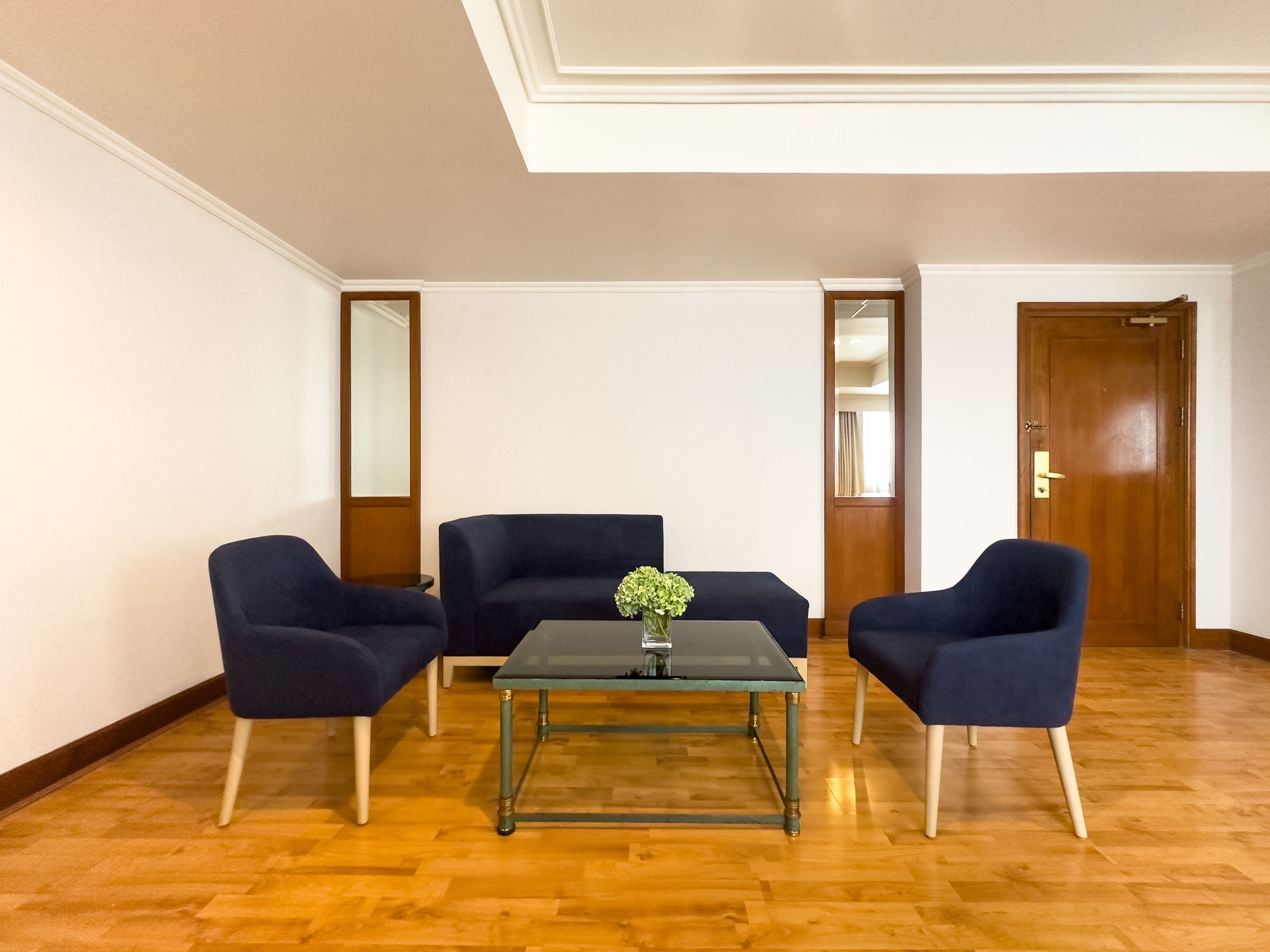 Une pièce avec un plancher en bois, deux fauteuils bleus, une table avec un plateau en verre sur laquelle est posé un vase de fleurs, et une porte en bois.