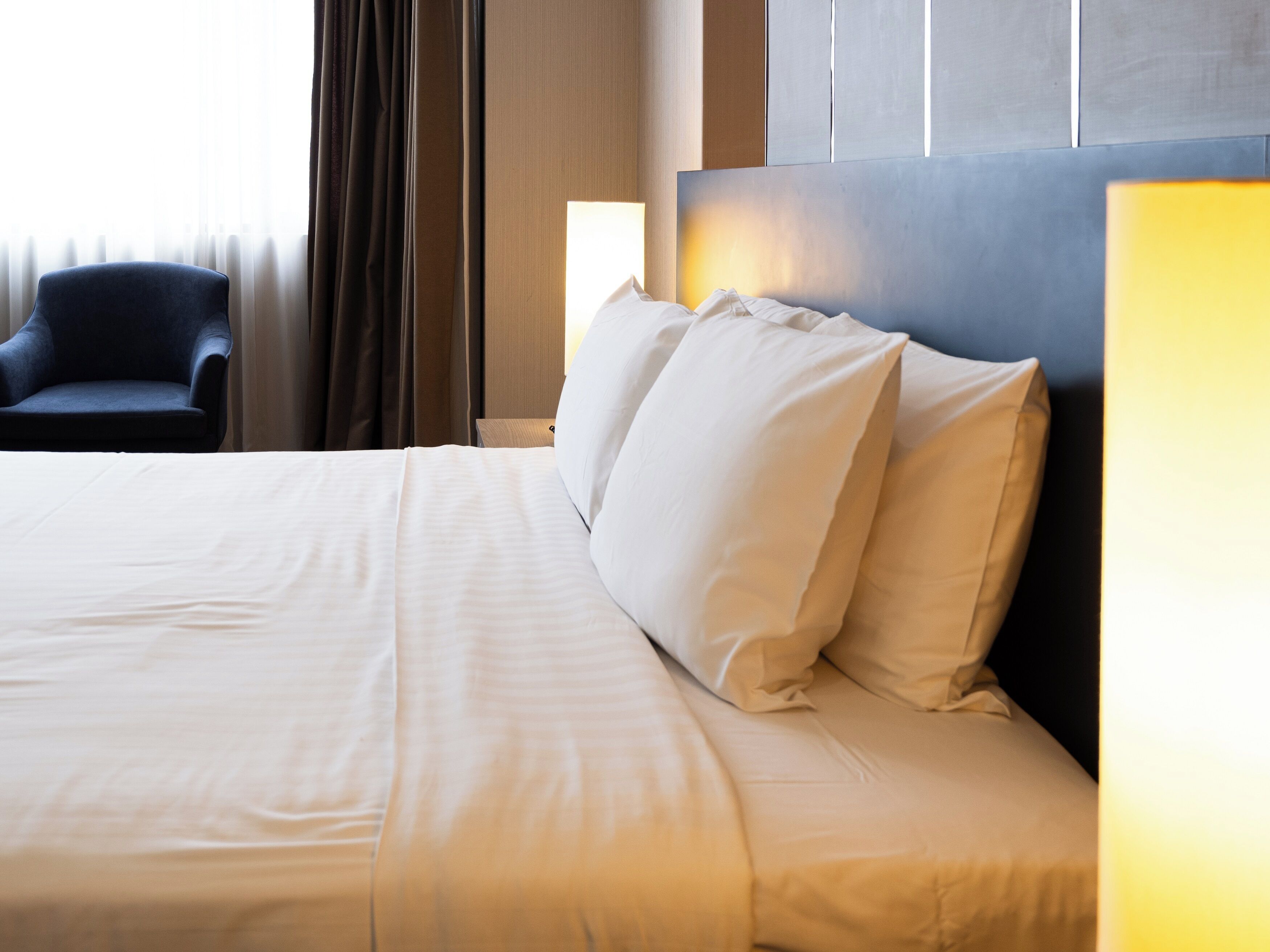 Una cama bien hecha con sábanas blancas y cabecera, un sillón oscuro y una ventana con cortinas.