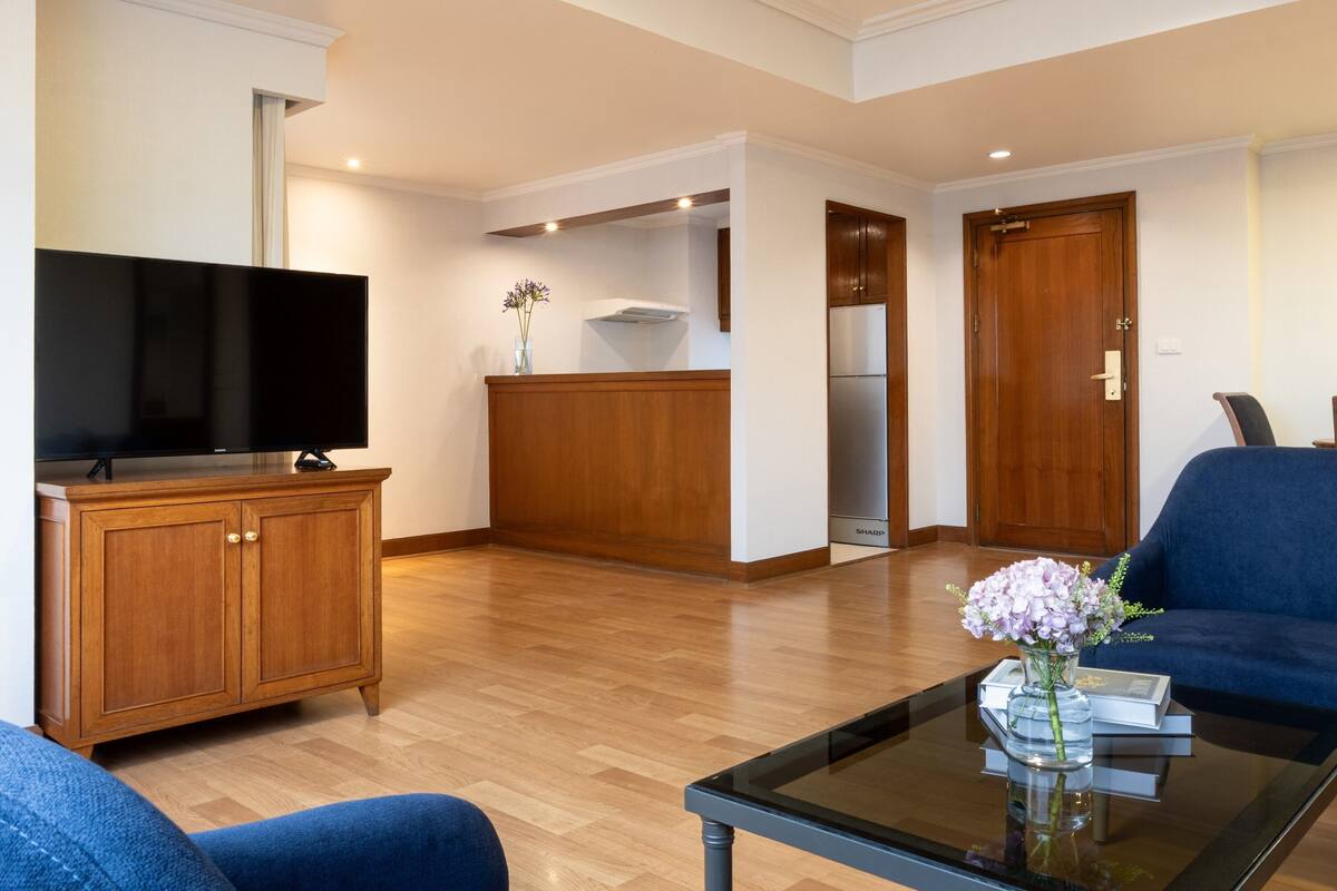 Una sala de estar moderna con un televisor de pantalla plana, muebles de madera, una mesa de centro de vidrio con un florero y un sofá azul.