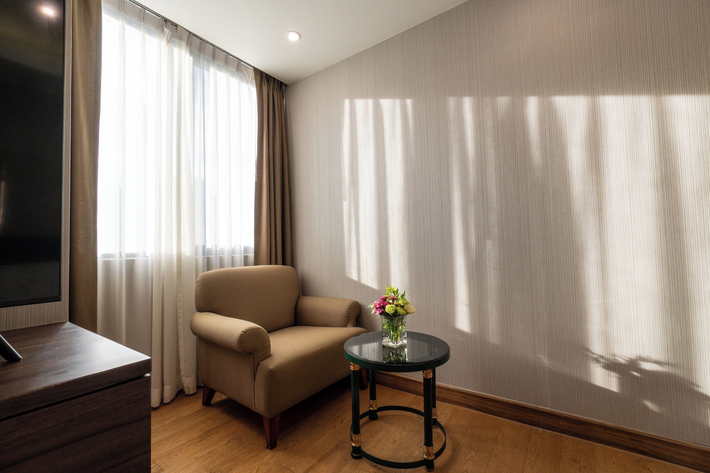 Una habitación con sofá, una pequeña mesa redonda con un jarrón de flores, televisor de pantalla plana y un amplio ventanal con cortinas.