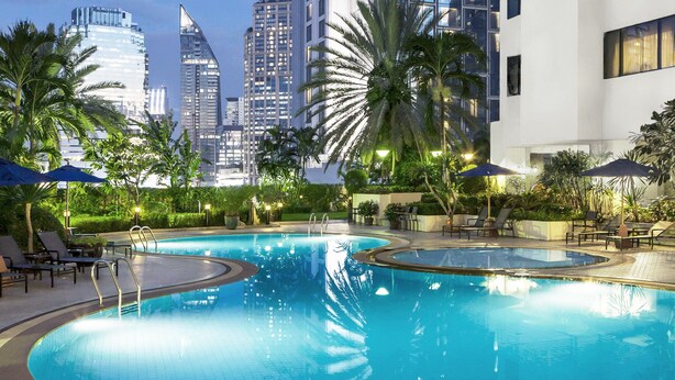 Piscina con sillas de descanso y sombrillas, rodeada de palmeras y edificios modernos al fondo.