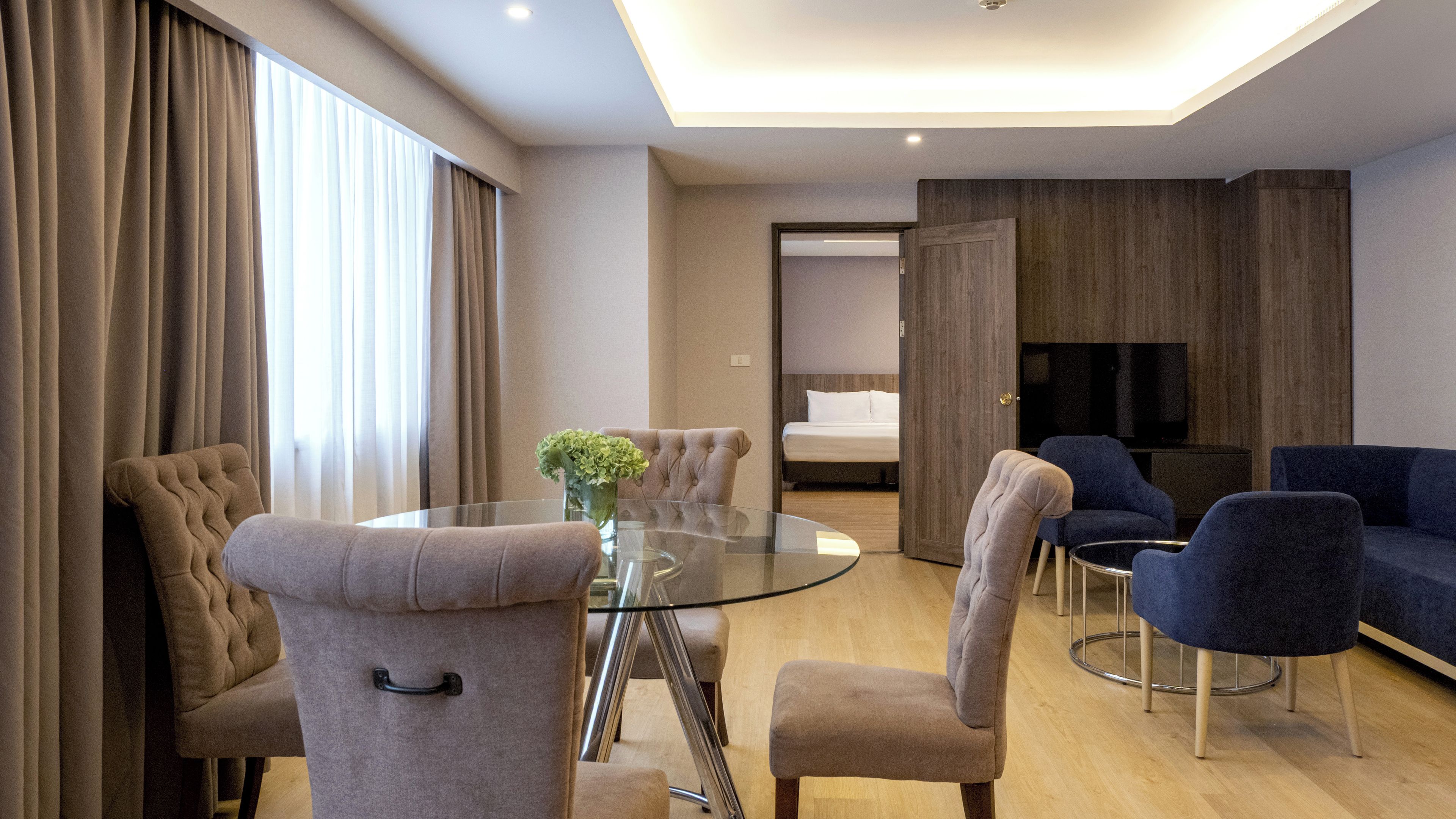 A modern hotel room with a round glass table, tufted armchairs, a sofa, and a bed with a wooden headboard.