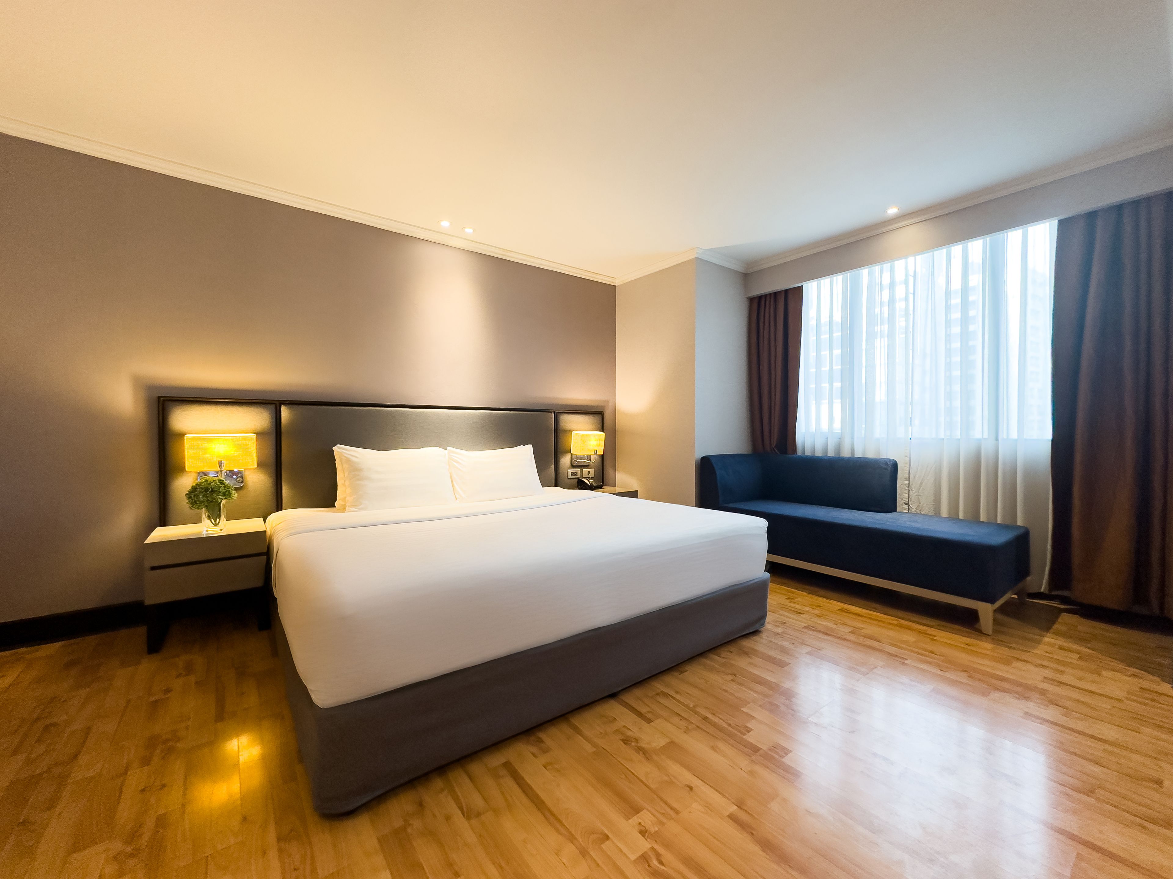 Een hotelkamer met een groot bed, bedlampen, een donkerblauwe bank en een groot raam met gordijnen.