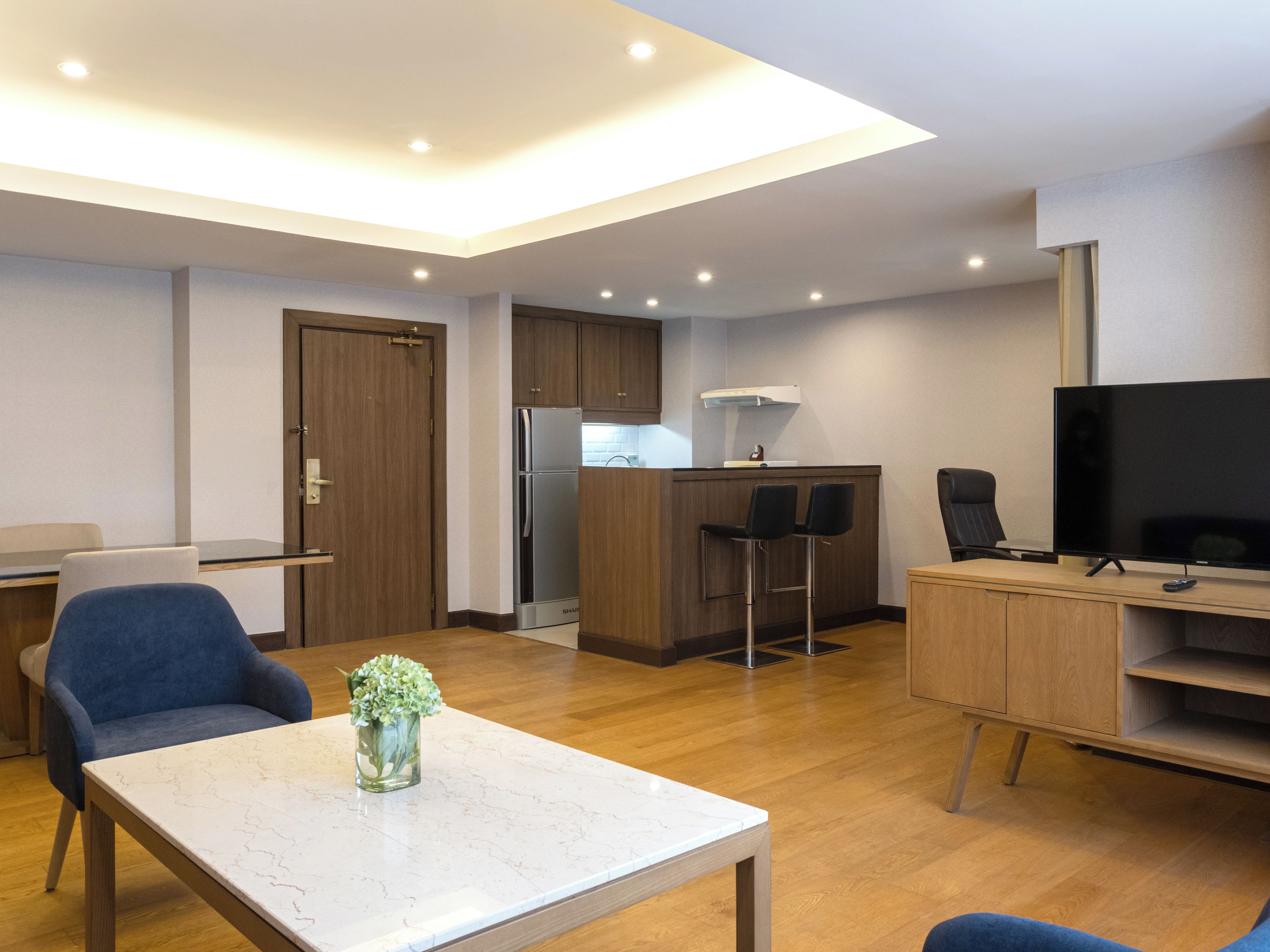 A modern living room with a marble coffee table, a TV, and a kitchen area with a refrigerator and bar stools.