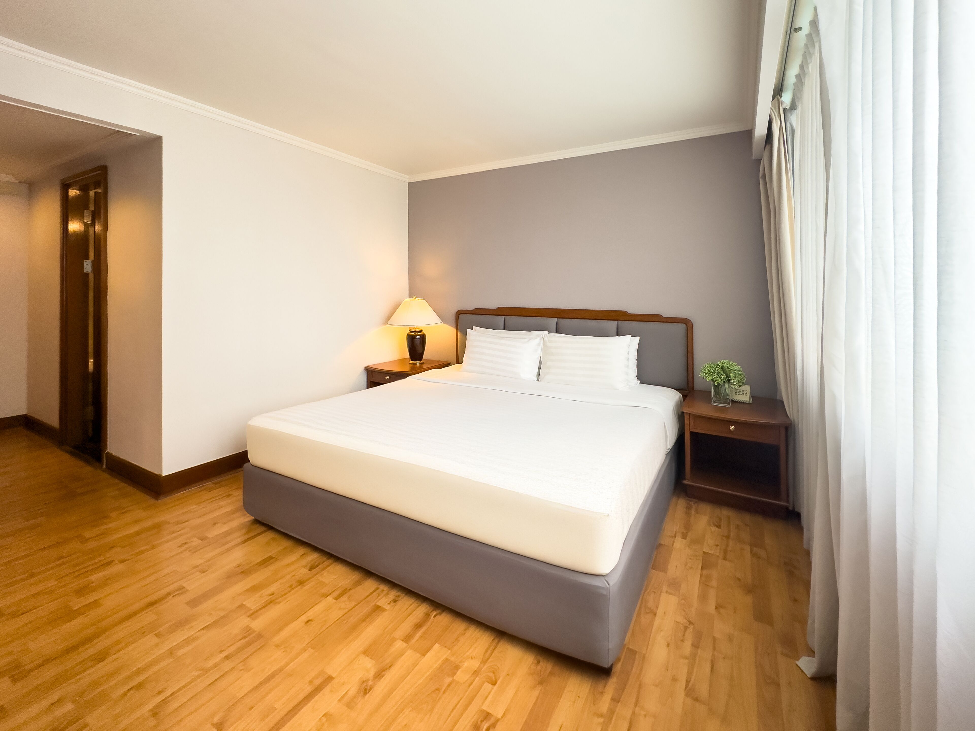 Een hotelkamer met een groot bed, houten vloer, een nachtkastje met een lamp en een kleine plant op het nachtkastje.