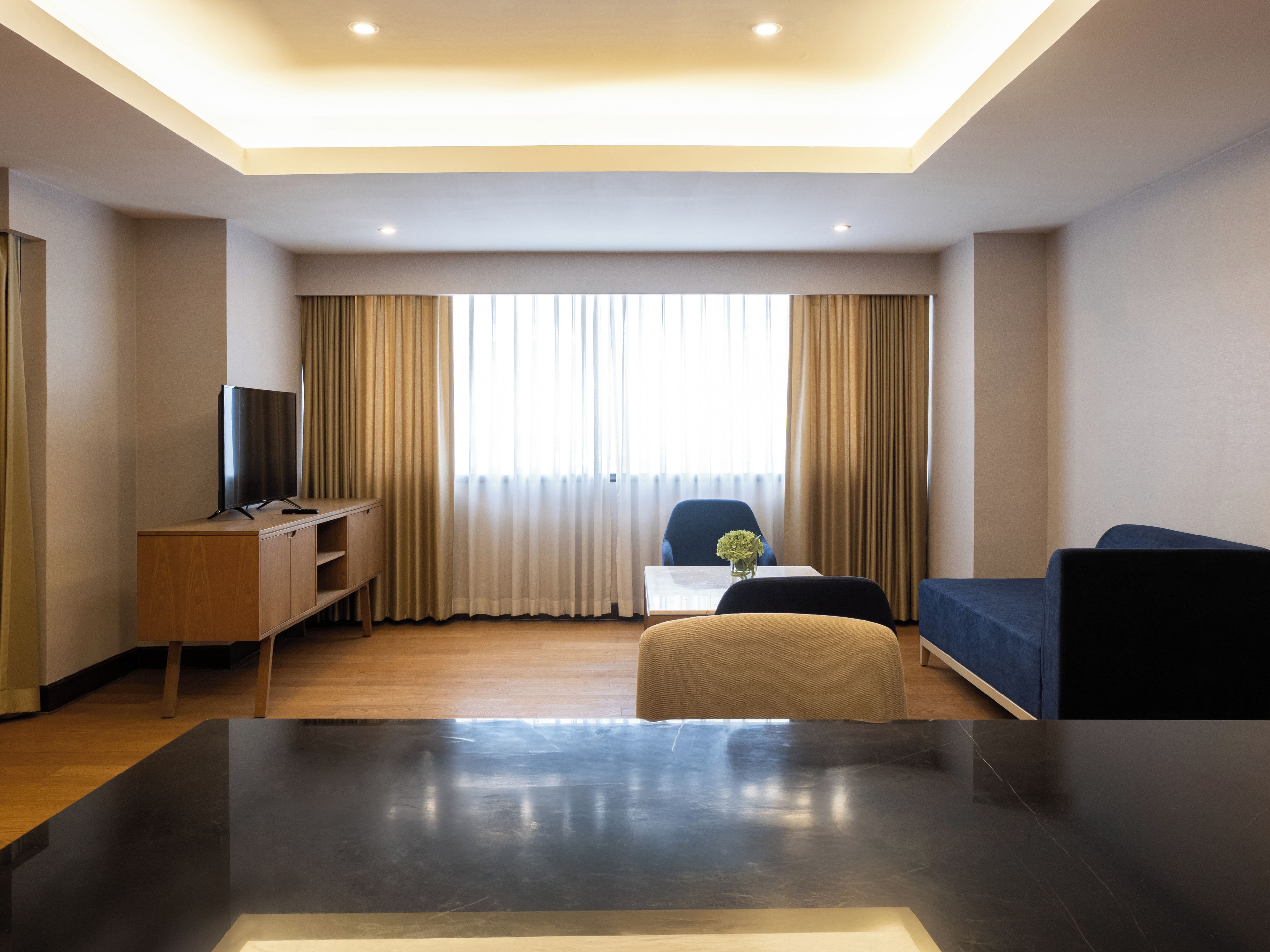 Ein modernes Hotelzimmer mit Fernseher auf einer Holzkommode, einer Couch, einem kleinen Tisch mit einer Vase und einem großen Fenster mit Vorhängen.