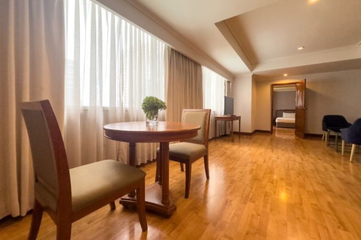 A hotel room with a wooden floor, a round dining table with chairs, a small side table, a bed with a canopy, and a large window with sheer curtains.