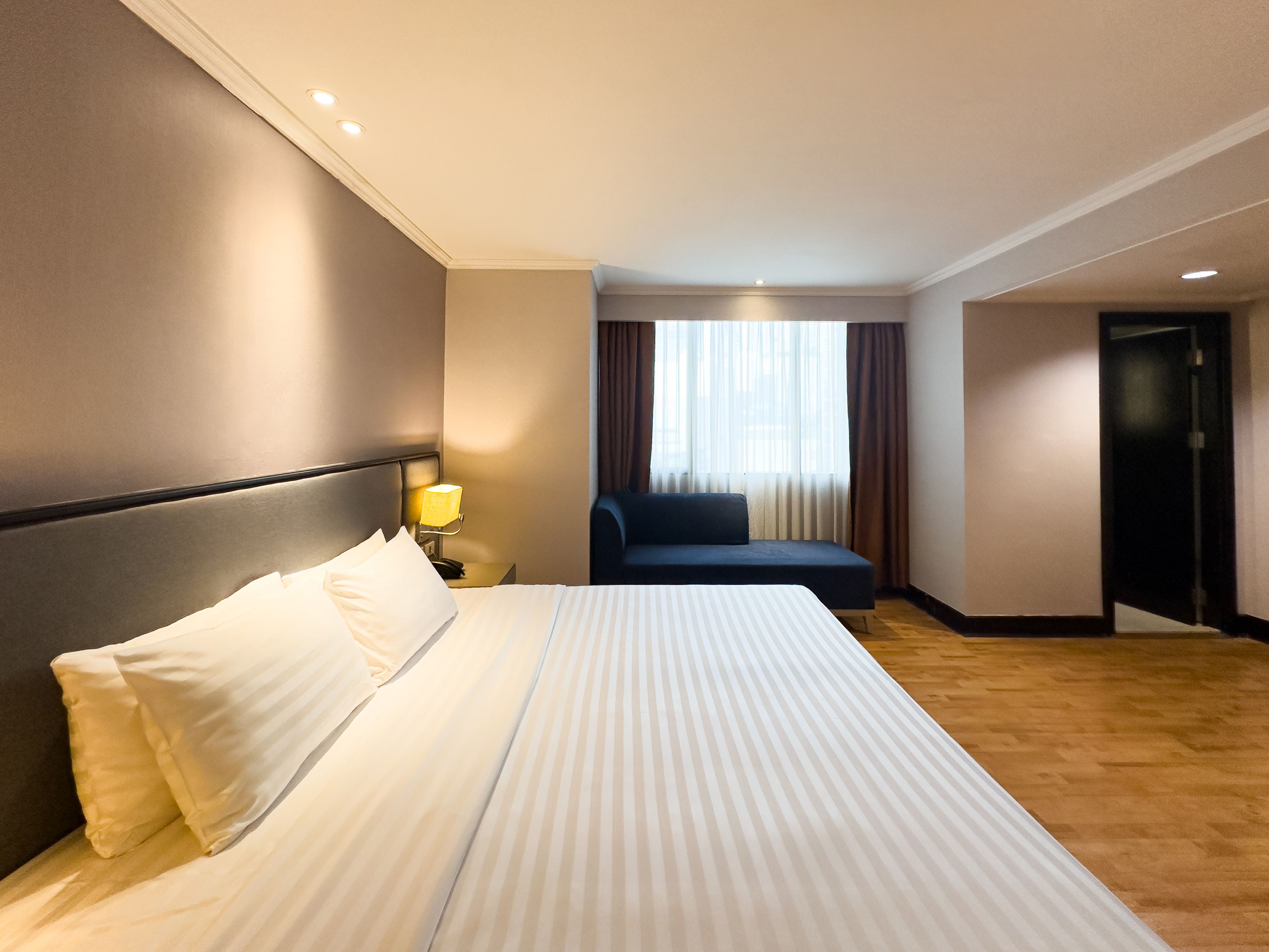 A hotel room with a large bed, a nightstand with a lamp, a seating area with a sofa, and a view of the outside through a window.
