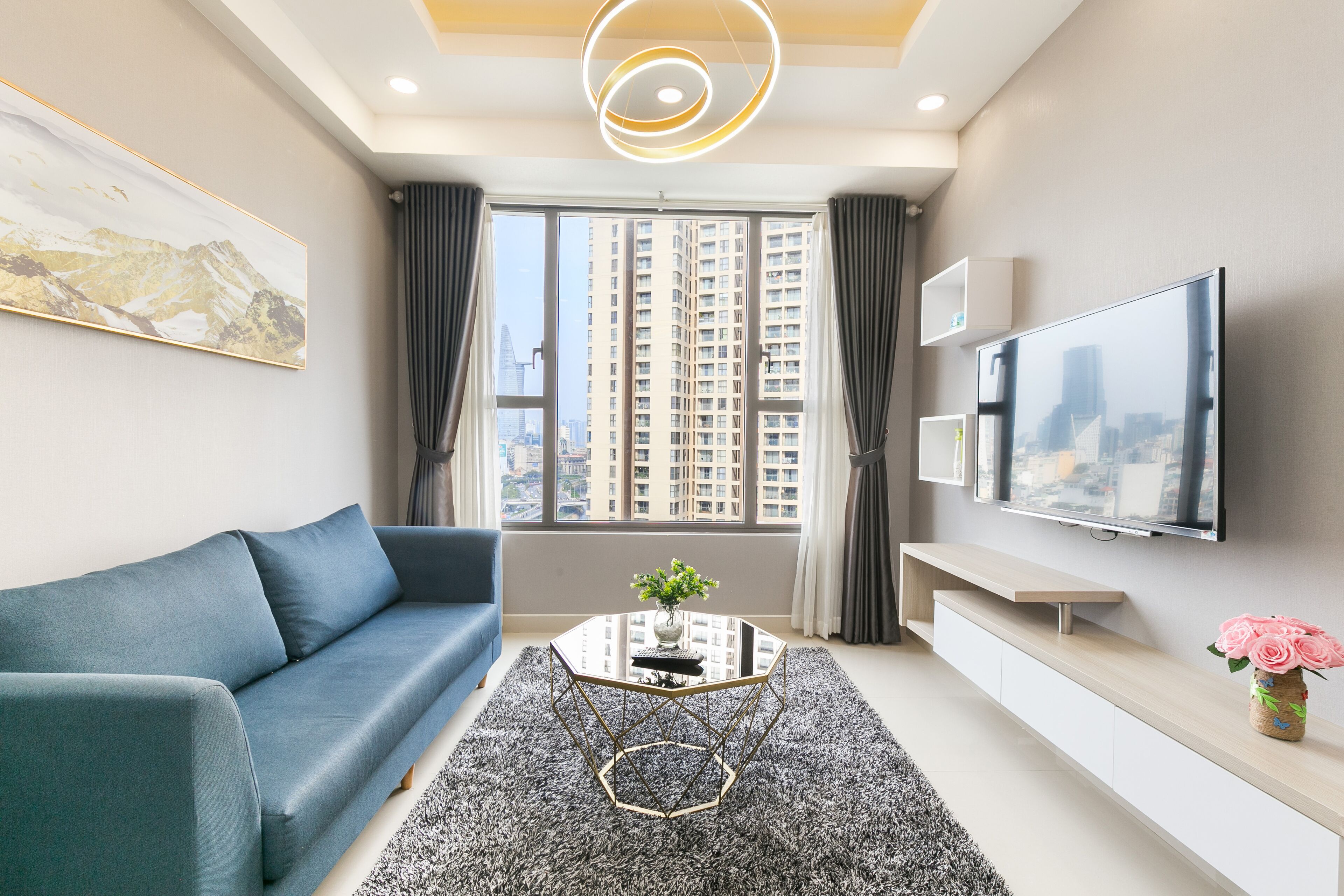 A modern living room with a sofa, a coffee table, a TV, and a view of tall buildings through large windows.